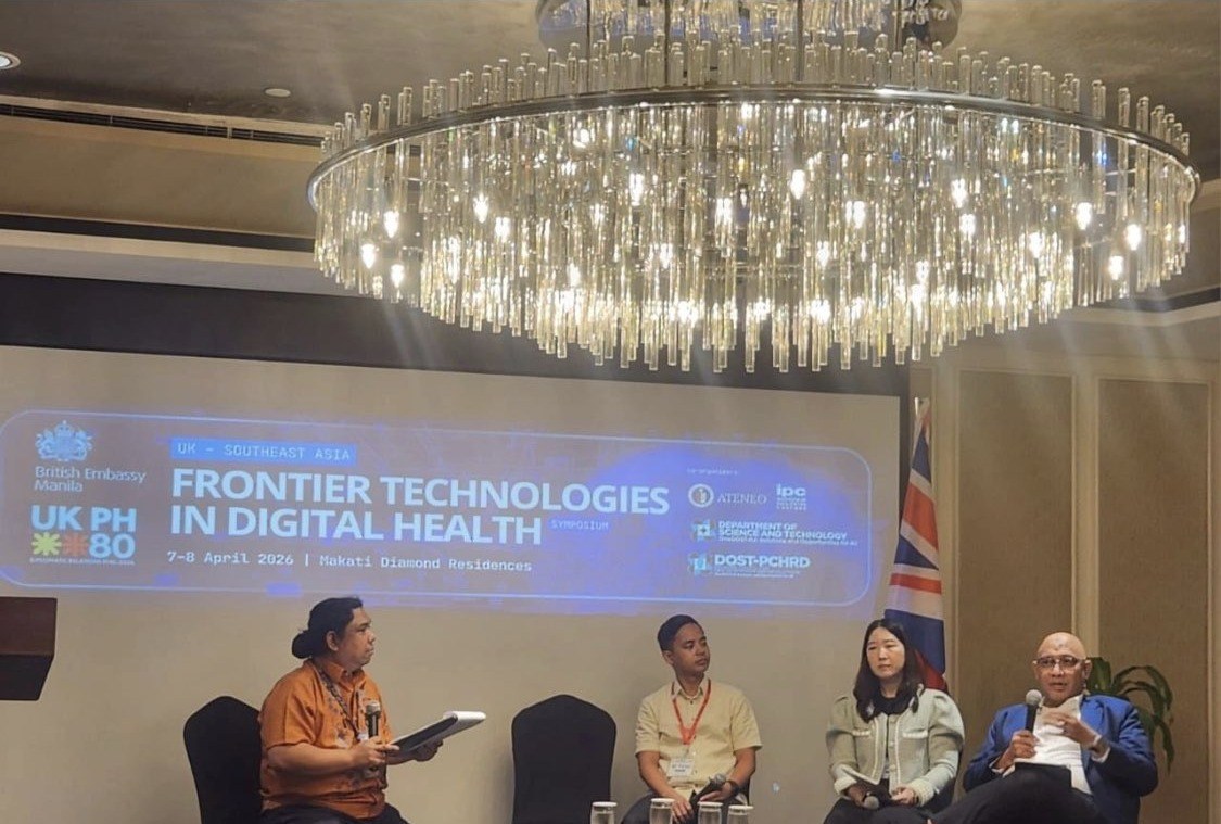 Reza Ali speaking from the stage at the UK-Southeast Asia Symposium on Frontier Technologies in Digital Health, seated with three other panellists beneath a screen showing the symposium title, its dates and Makati Diamond Residences as the venue.