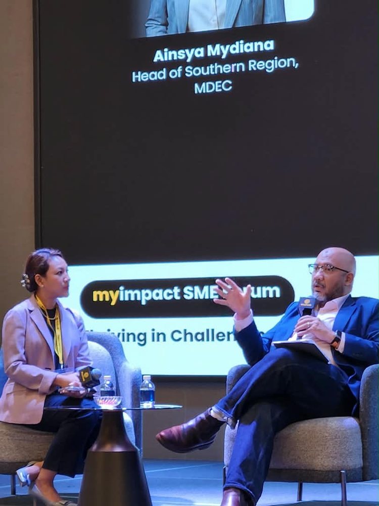 Reza Ali mid-sentence, microphone in hand, seated on stage beside Ainsya Mydiana of MDEC during a fireside session at the Maybank myimpact SME Forum in Johor Bahru.