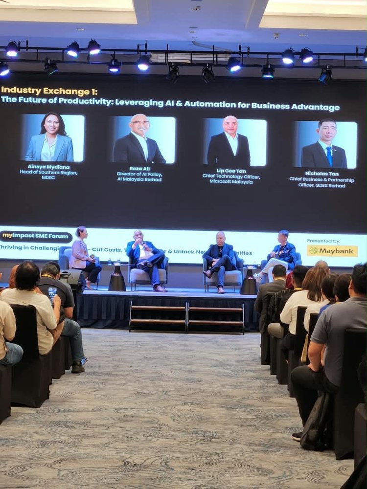 The Industry Exchange panel on stage beneath a screen showing the session title and the four panellists' portraits: Ainsya Mydiana of MDEC, Reza Ali, Lip Gee Tan of Microsoft Malaysia and Nicholas Tan of GDEX. The audience is seated in rows in the foreground.