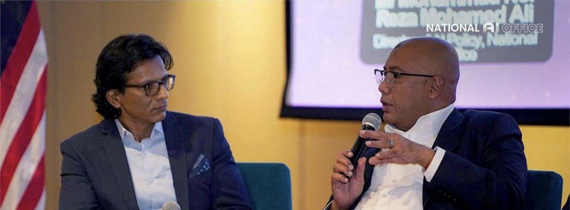 Reza Ali speaking with a handheld microphone at WASIIM 2026, in a dark blazer over a white shirt, seated opposite another participant, with the event screen behind him carrying his name, his title and his office.