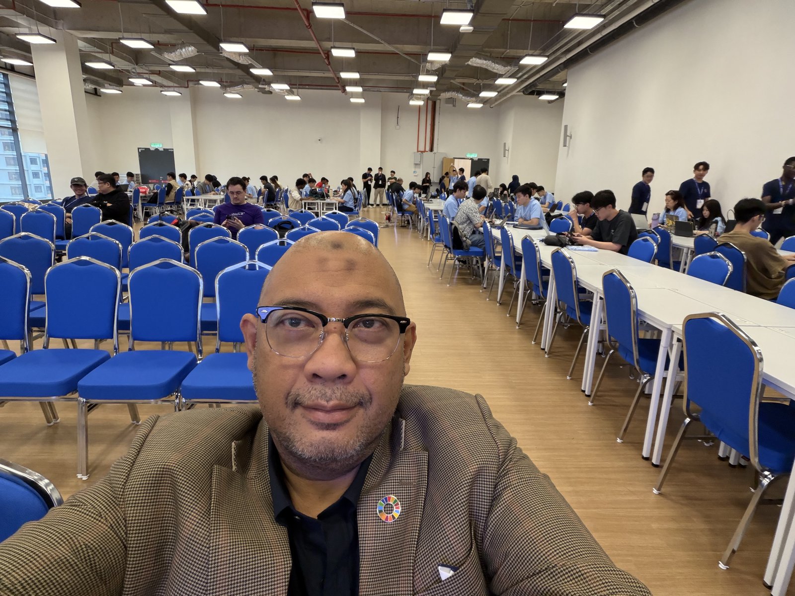 Reza Ali at the Codex Hackathon with participants working at laptops in the hall behind him