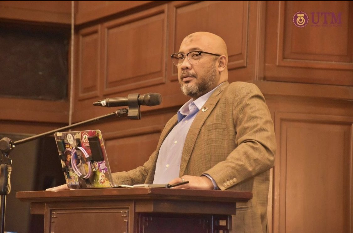 Reza Ali speaking from a wooden lectern at ISMI2026, holding a pen, with a laptop in front of him, in a wood-panelled hall.